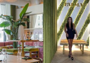 L'image montre un interieur elegant du restaurant Palma a Montreal, avec un decor moderne et chaleureux. A gauche, on voit une salle de restaurant lumineuse avec des plantes exotiques, des tables en bois bien dressees avec des verres et des couverts, et des banquettes en velours rose. A droite, une femme portant une robe noire chic et des talons hauts pose devant un mur vegetal eclaire avec le logo "Palma" au-dessus